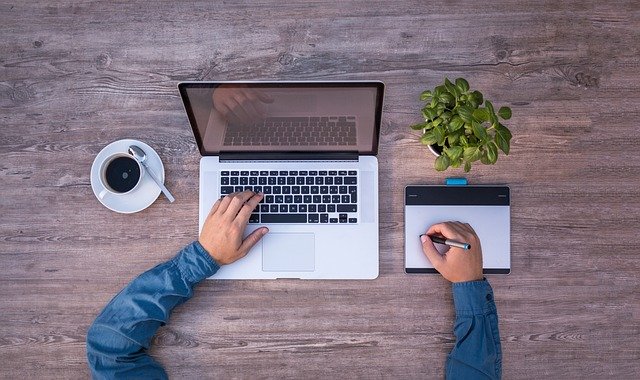 Na imagem, uma vista aérea em close, de um homem trajando camisa azul, trabalhando em um computador e fazendo uma assinatura eletrônica em um tablet gráfico. Sobre a mesa, além do computador, há uma xícara de café e uma planta